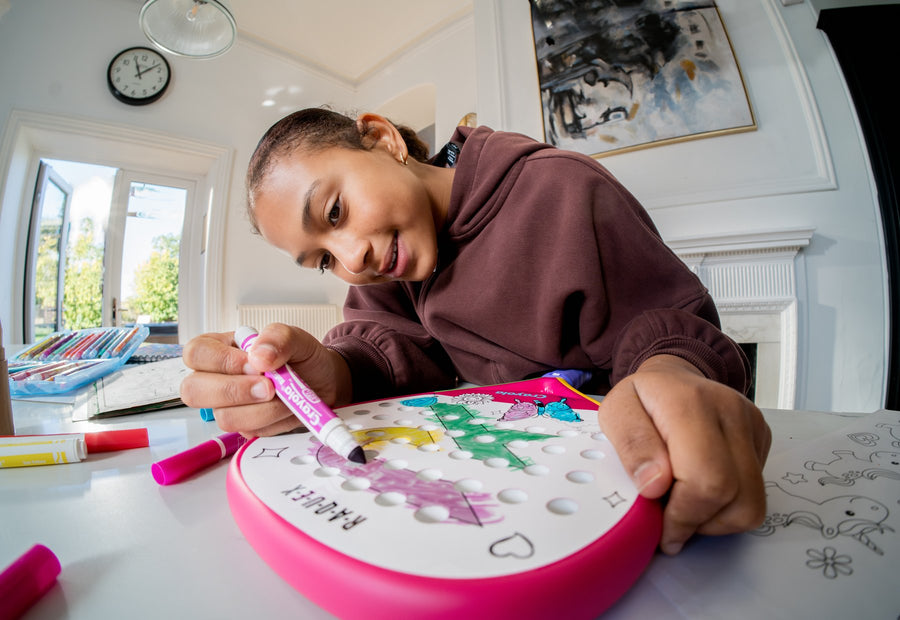 A girl leaning over a table colouring the face of a pink Raquex x Crayola racquet with a green marker