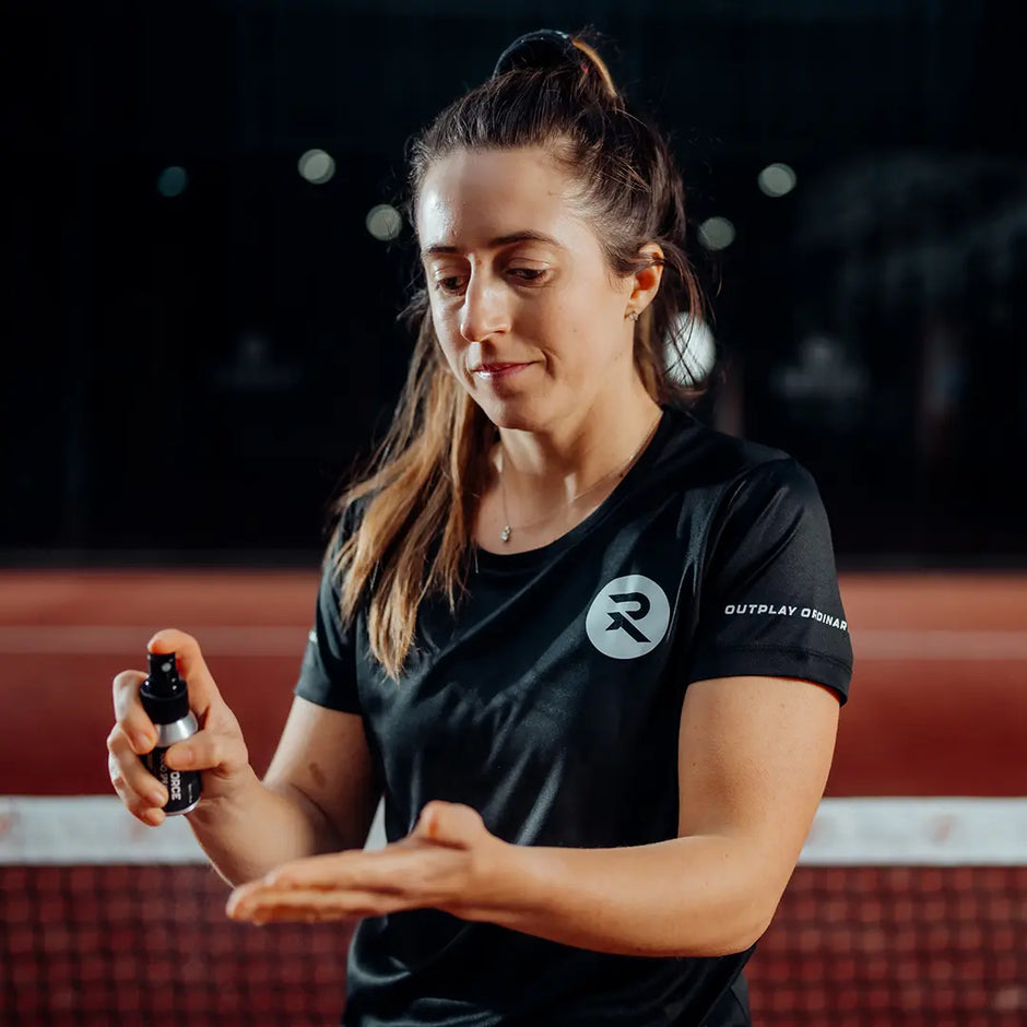 A woman in a black athletic t-shirt with a white circular logo and 'OUTPLAY ORDINARY' text on the sleeve, holding a small spray bottle over her hand in a dark, indoor sports environment.