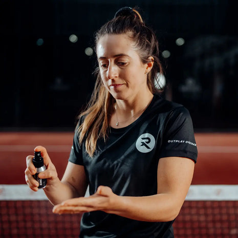 A woman in a black athletic t-shirt with a white circular logo and 'OUTPLAY ORDINARY' text on the sleeve, holding a small spray bottle over her hand in a dark, indoor sports environment.