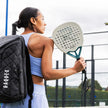 Woman holding a paddle racquet with a raquex sports bag on her back, standing on a tennis court.