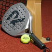 Raquex Evion Match padel racquet in silver resting against a padel net post, with a Raquex Padel Elite Pace ball on the red court