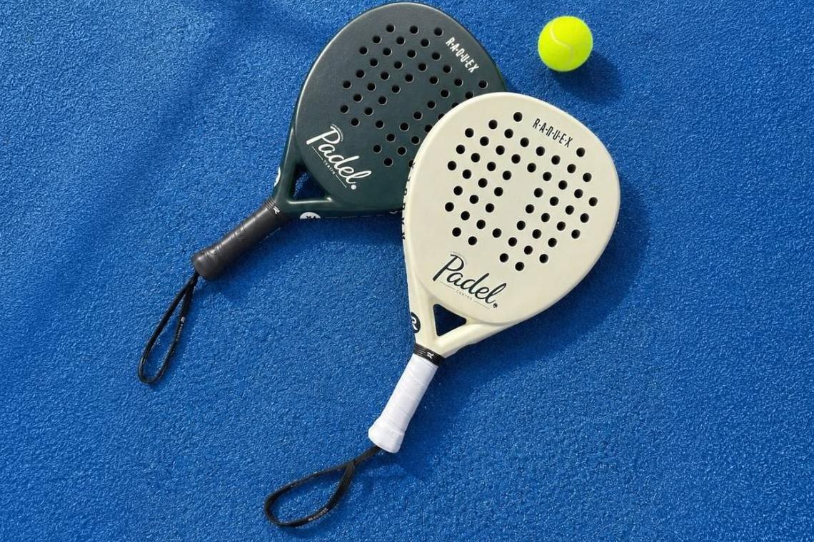 Two Raquex padel racquets co-branded with the Padel Centre logo, one dark green and one cream, laid on a blue court beside a ball