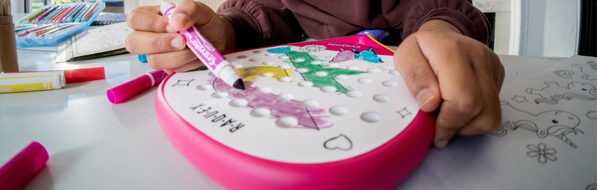 Close crop of a child colouring the white face of a pink Raquex x Crayola padel racquet with a Crayola marker, more markers and a colouring sheet on the table