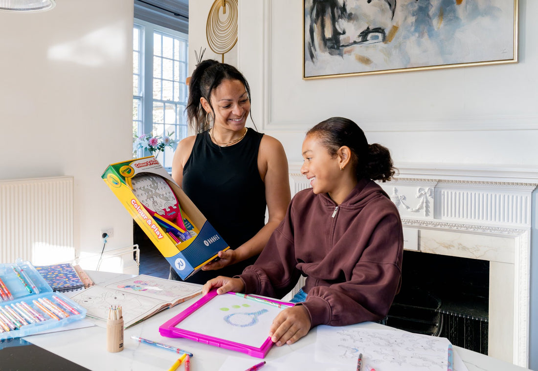 A mother holding up the boxed Raquex x Crayola Colour and Play pack while her daughter colours a racquet at the table