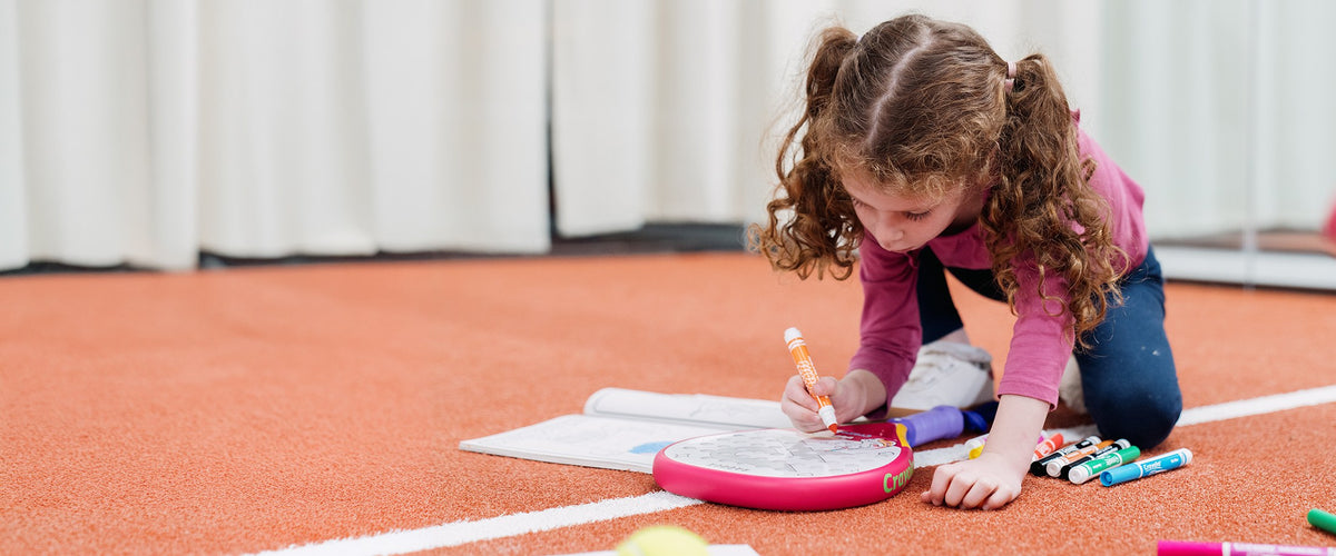 A young girl kneeling on an indoor padel court, colouring the face of a pink Raquex x Crayola junior racquet with an orange marker