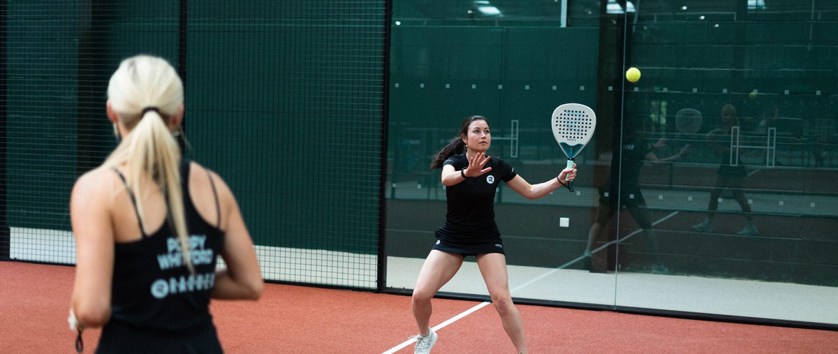 Two women playing padel on an indoor glass-walled court, wearing Raquex kit