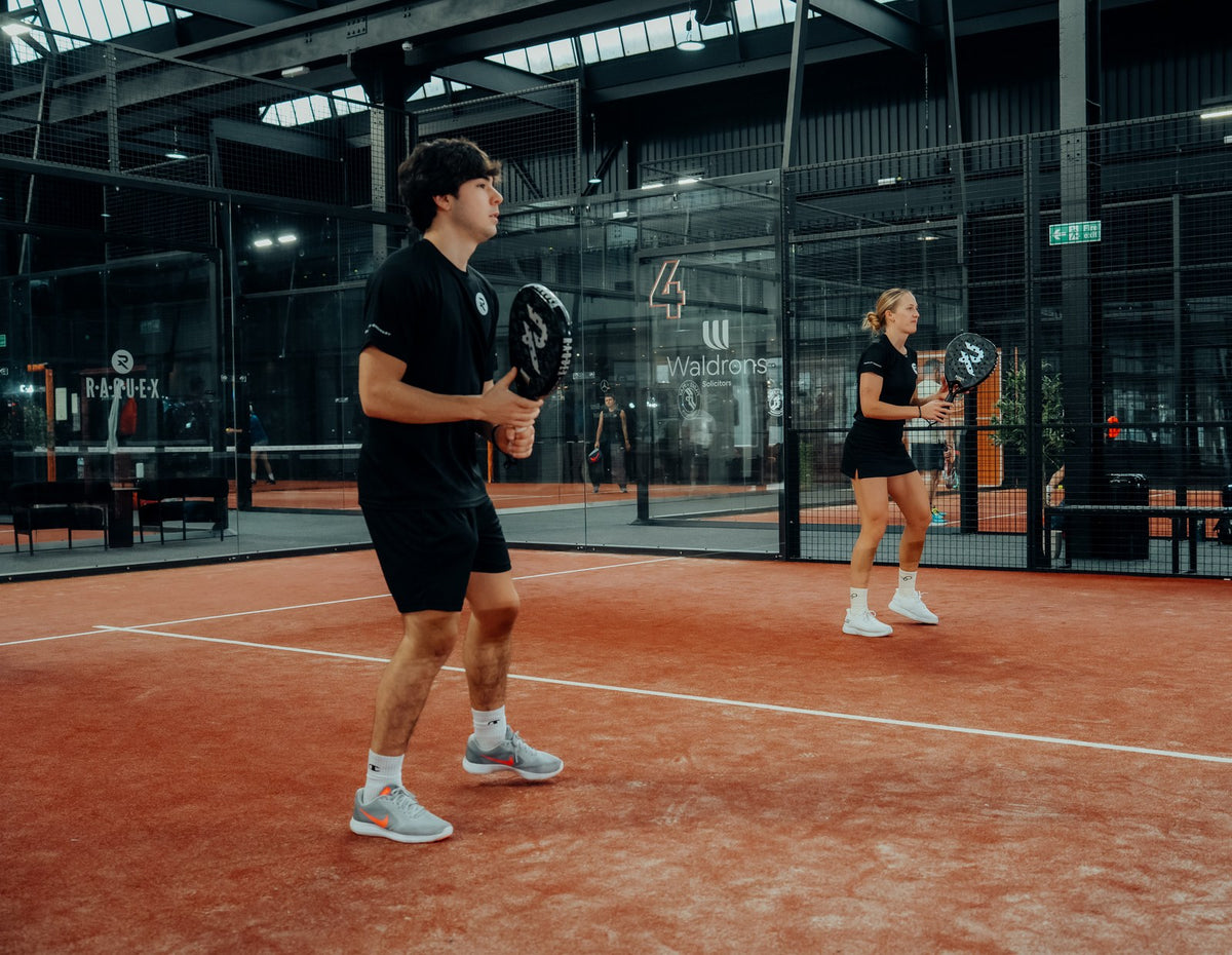 Two players mid point on an indoor clay padel court at a Raquex partner club, with Raquex boards on the glass