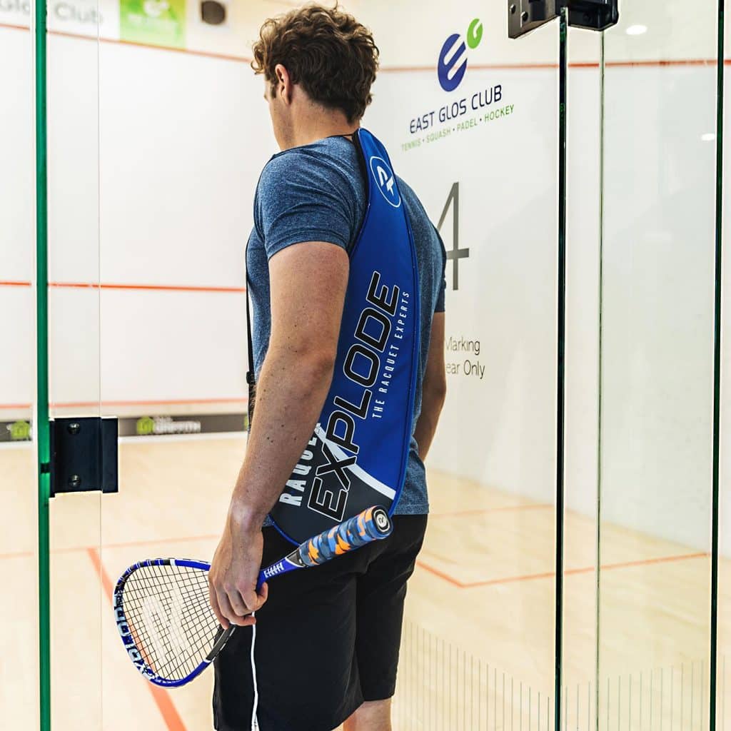 Man with a blue squash racquet with a blue and orange racquet grip, entering a squash court